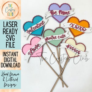 May include: A collection of colorful heart-shaped decorations on wooden sticks. Each heart has a different color and a banner with a Valentine's Day message like "Love" or "Kisses". The image also includes text: "LASER READY SVG FILE".