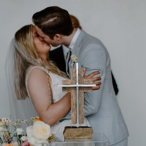 May include: A couple kisses during their wedding ceremony, with a wooden cross with silver accents in the foreground.