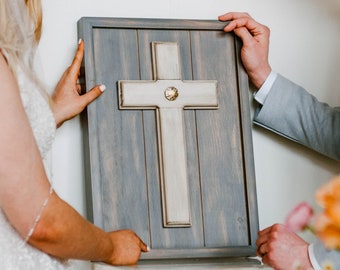 May include: A wooden cross with a light-colored center and a decorative gem. The cross is mounted on a gray wooden panel with a dark gray frame. The cross is being held by two people.