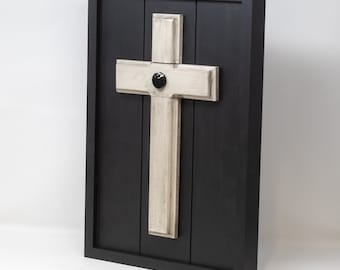 May include: A white wooden cross with a black circle in the center is mounted on a black wood background. The cross is framed in a black wood frame.