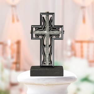 May include: A decorative cross with a black frame and a silver-coloured centre, set on a dark grey rectangular base. The cross features ornate detailing and is displayed on a white pedestal.