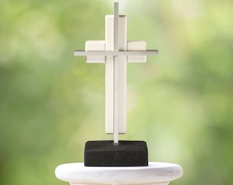 May include: A white wooden cross with a silver metal frame sits on a black base. The cross is on a white pedestal against a green blurred background.