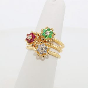 Puede incluir: Tres anillos de oro, cada uno con un diseño floral. Un anillo presenta piedras preciosas rojas, otro verdes y el tercero tiene piedras preciosas claras. Los anillos se exhiben sobre una superficie blanca.