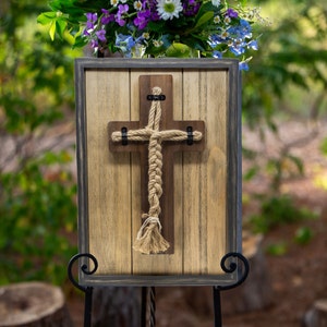 Könnte beinhalten: Ein Holzkreuz mit einem Seildesign wird in einem rustikalen Rahmen auf einem schwarzen Staffelei ausgestellt. Der Rahmen ist aus Holz mit einer abgenutzten Oberfläche. Das Kreuz ist hellbraun und das Seil ist naturfarben. Der Hintergrund ist verschwommen und unscharf.