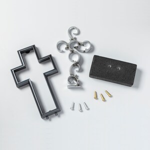 K&ouml;nnte beinhalten: Ein schwarzes Metallkreuz, ein silbernes Metall-Dekorationsst&uuml;ck, eine schwarze rechteckige Basis und sechs kleine Metall-Stifte. Die Stifte sind in zwei Gruppen zu je drei, eine Gruppe ist golden und die andere ist silber.