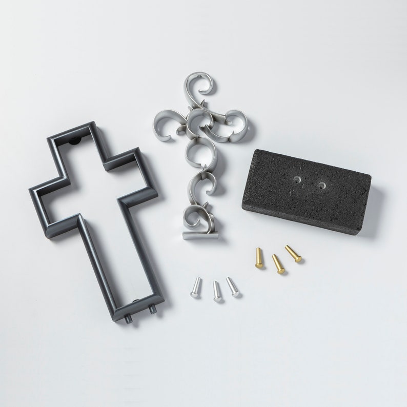 K&ouml;nnte beinhalten: Ein schwarzes Metallkreuz, ein silbernes Metall-Dekorationsst&uuml;ck, eine schwarze rechteckige Basis und sechs kleine Metall-Stifte. Die Stifte sind in zwei Gruppen zu je drei, eine Gruppe ist golden und die andere ist silber.