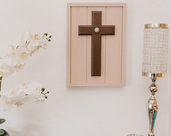 May include: A brown wooden cross with a white gem in the center, mounted on a light pink wood frame.
