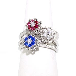 May include: A silver-toned ring featuring three flower-shaped clusters. Each cluster is set with small, sparkling stones in red, blue, and clear white. The band has a textured, wavy surface, and the floral settings are arranged in a tiered, overlapping design.