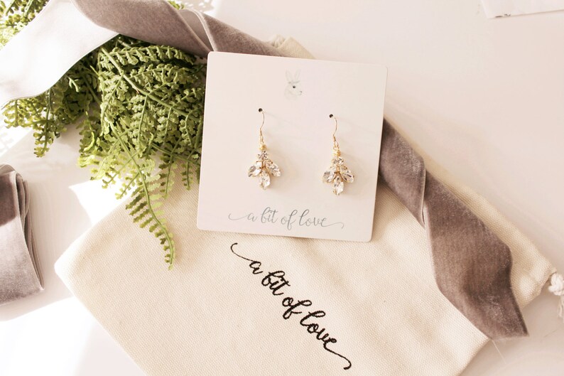 Op de afbeelding: Een paar gouden oorhangers met heldere kristallen accenten. De oorbellen worden gepresenteerd op een witte kaart met de tekst "a bit of love".
