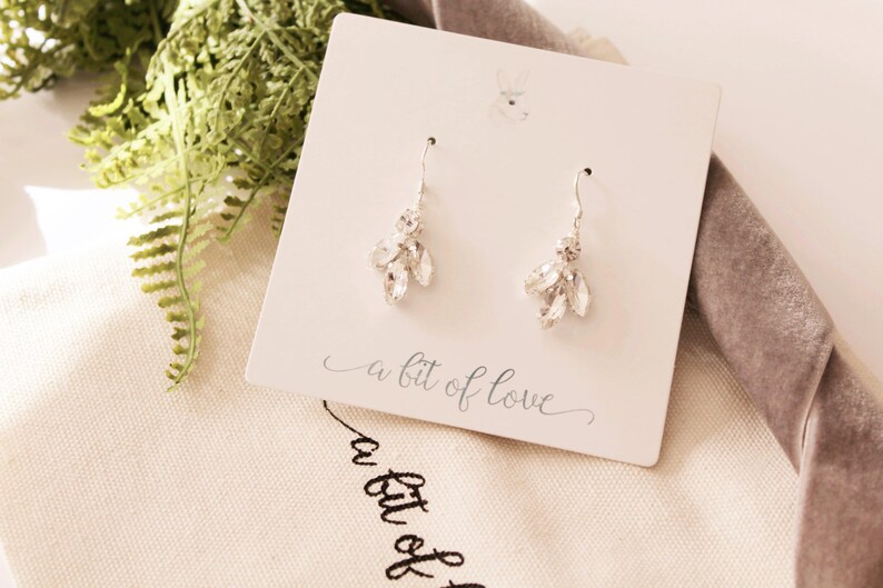 Op de afbeelding: Een paar zilveren oorhangers met heldere kristallen accenten. De oorbellen bevinden zich op een witte kaart met de tekst "a bit of love".
