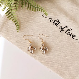 Op de afbeelding: Een paar gouden oorhangers met heldere kristallen. De oorbellen liggen op een witte stoffen achtergrond met de tekst "a bit of love" erop geborduurd.