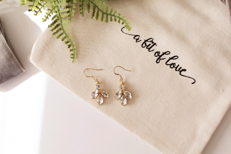 Op de afbeelding: Een paar gouden oorhangers met heldere kristallen. De oorbellen liggen op een witte stoffen achtergrond met de tekst "a bit of love" erop geborduurd.