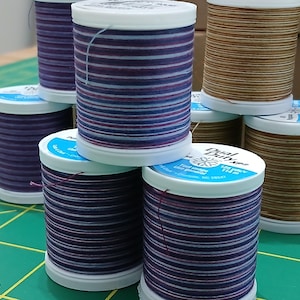 May include: A collection of spools of thread in various colors, including purple, brown, and blue. The spools are stacked on a green cutting mat. The thread spools have white tops with blue labels that read "Dual Duty XP".