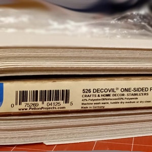 May include: A rolled fabric, labelled as Pellon 526 Decovil One-Sided Fusible Stabilizer. The label indicates a width of 43 cm. The fabric is off-white and intended for crafts and home decor.