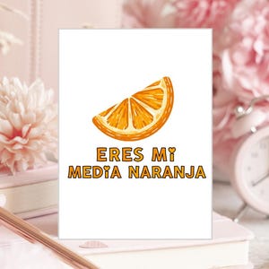 May include: A white greeting card featuring a graphic illustration of a bright orange citrus slice at the top. Below the orange slice, the text ERES MI MEDIA NARANJA is printed in bold, orange, sans-serif capital letters with small heart accents.
