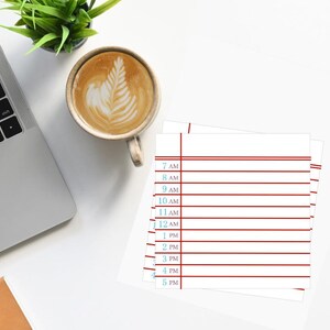 May include: A square white notepad featuring a daily schedule layout with horizontal red lines and blue text indicating times from 7 AM to 5 PM. The notepad is positioned on a flat surface next to a laptop, a cup of coffee, and a small green plant.