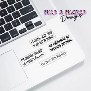 May include: A collection of white stickers with black text on a laptop. The stickers feature phrases in English and Spanish, including "I Belong aqui, auá y en TODAS PARTES" and "Plot Twist: We're Still Here". The laptop is silver and has a black keyboard.
