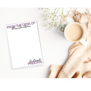 May include: A white notepad with the text "FROM THE DESK OF Mrs. Jackson" and "School Principal" in a script font. A white mug filled with a light brown liquid sits next to the notepad, along with a beige cloth and white flowers.