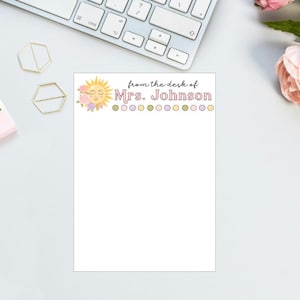 May include: A personalized notepad with the text "from the desk of Mrs. Johnson." The top of the notepad features a sun illustration with floral accents and a row of colorful dots. A keyboard, gold paper clips, and flowers are in the background.