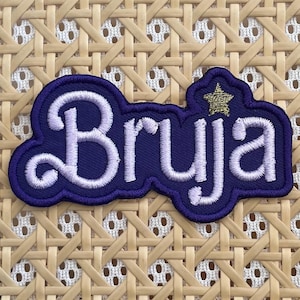 May include: Purple embroidered patch with the word "Bruja" in white lettering and a gold star above the word.