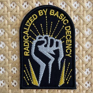 May include: Black and gold embroidered patch with a raised fist and the text "Radicalized by Basic Decency".