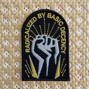 May include: A black embroidered patch with an arched top featuring a silver raised fist graphic in the center. The text RADICALIZED BY BASIC DECENCY is stitched in yellow thread in an arc above the fist, with yellow sunburst rays surrounding it.