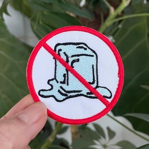 Peut inclure: Un écusson rond blanc avec une bordure rouge représentant un glaçon de dessin animé barré d'une ligne rouge. Le glaçon est bleu clair et en train de fondre. L'écusson est présenté sur un fond de feuilles vertes.