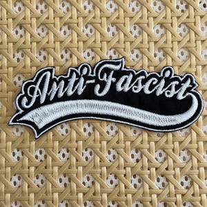 Peut inclure: Écusson brodé avec les mots "Anti-Fasciste" en écriture blanche sur fond noir. Le patch a une bordure blanche et est de forme rectangulaire. Le fond est un motif tissé brun clair.