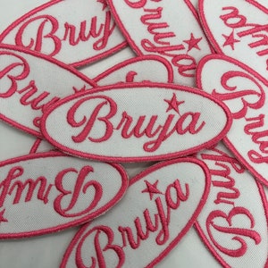 May include: A pile of white oval patches with pink embroidered lettering that says "Bruja" with a star.