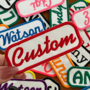 May include: A white iron-on patch with a red border and the word "Custom" in red cursive lettering.
