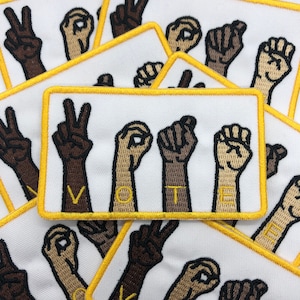 May include: Multiple white patches with yellow borders, each featuring a different hand sign spelling out the word "VOTE" in American Sign Language. The patches are made of fabric and have a stitched appearance.