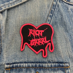 May include: A black heart-shaped patch with pink dripping edges. The patch has the words "Riot Grrrl" written in pink inside the heart.