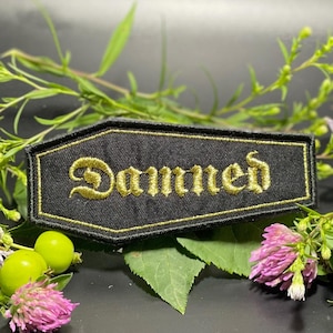Peut inclure: Écusson noir en forme de cercueil avec le texte brodé en or "Damned". L'écusson a une bordure dorée et est entouré de feuillage vert, de trèfles roses et de baies vertes. L'écusson est probablement destiné aux vêtements ou aux accessoires.