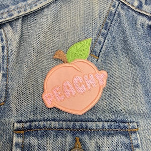 May include: A pink peach-shaped patch with the word "Peachy" embroidered in pink on a denim jacket.