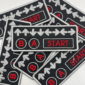 May include: A set of black and white embroidered patches with red accents. The patches feature a variety of gaming controller buttons, including "A", "B", "Start", and directional arrows.
