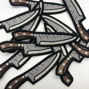 May include: A pile of embroidered iron-on patches featuring a cartoon knife design. The knives are gray with brown handles and white stitching.
