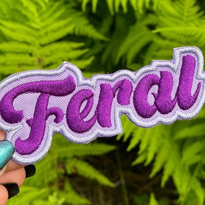 May include: Purple "Feral" embroidered patch with a light purple background. The patch is made of fabric and has a stitched edge.