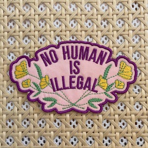 Peut inclure: Un patch en tissu rose avec des coutures violettes et des fleurs jaunes. Le patch dit "No Human Is Illegal" en lettres violettes.