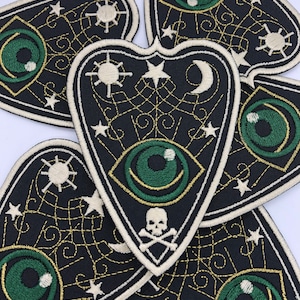 May include: Five black and white embroidered patches in the shape of a heart with a green eye, stars, a crescent moon, a sun, and a skull and crossbones. The patches have a gold thread outline and are on a white background.