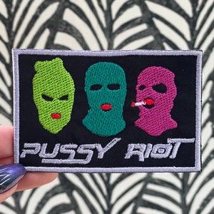 Peut inclure: Écusson brodé avec trois visages cagoulés en vert, turquoise et rose, chacun avec des lèvres rouges. Le texte "PUSSY RIOT" est brodé en blanc sous les visages sur un fond noir, encadré d'une bordure blanche.