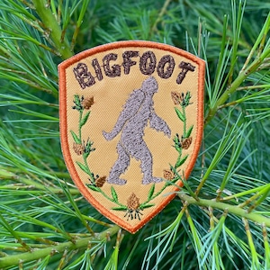 May include: A shield-shaped embroidered patch with a tan background and a burnt orange border. The patch features a dark brown embroidered silhouette of Bigfoot in the center, the text BIGFOOT in brown, and green pine branches with brown cones.