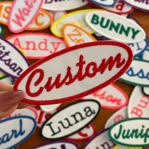 May include: A close-up of a white oval patch with a red embroidered word "Custom" on it. The patch is surrounded by other oval patches with various names and colors.