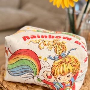 Może przedstawiać: Biały ceramiczny kubek z czarnym uchwytem i napisem "But First, Coffee" pogrubioną, czarną czcionką. Kubek jest ustawiony na rozmytym tle, sugerującym przytulne otoczenie.