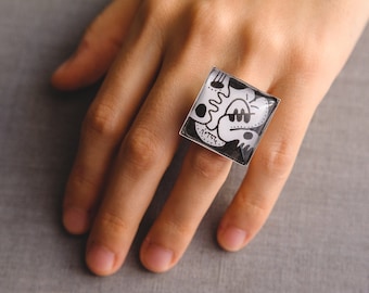 May include: A square ring with a black and white abstract design. The ring is made of resin and is set on a silver band.