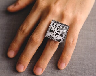 May include: A square-shaped ring with a black and white abstract design. The ring is made of clear resin and has a glossy finish.