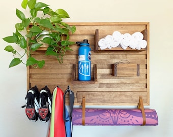 May include: A wooden wall organizer with hooks for hanging items, a shelf for storing towels, and a space for a water bottle. There is a yoga mat hanging from the bottom of the organizer, and a plant is hanging from the top.