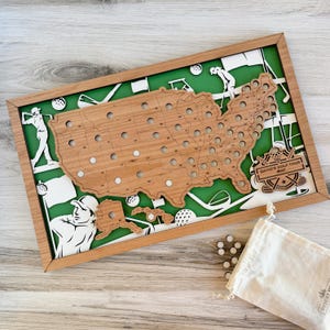 May include: A wooden golf tour map of the United States with a green background and a wooden frame. The map has holes for marking locations and includes golf-themed cutouts. A small bag of white markers is included.