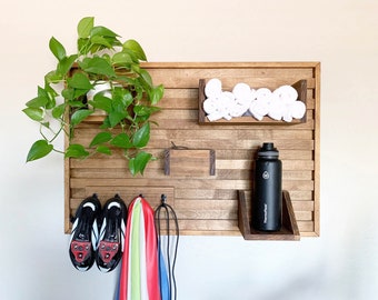 May include: A wooden wall organizer with hooks, shelves, and a small cubby. It features a black water bottle, white towels, and a green plant. There are also two black cycling shoes and a multi-colored towel hanging on the hooks.