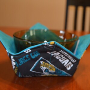 May include: A black and teal bowl cozy with the Jacksonville Jaguars logo and text. The cozy is folded to hold a glass bowl. The interior is teal, and the exterior features the team's logo and colors. The cozy is on a wooden table.