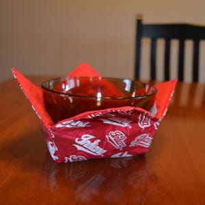 May include: A clear glass bowl sits inside a red and white fabric bowl cozy. The cozy is patterned with white text and logos on a red background. The bowl and cozy are on a wooden table.